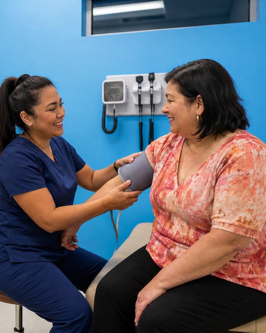 A nurse checks an older woman's blood pressure in an exam room with a bright blue wall, the two of them smiling at each other